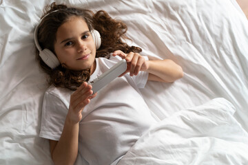 beautiful teenage girl looks at smartphone screen with headphones in bed in the morning. Happy adult child wakes up and listens to music, plays games, studies with lessons on the phone