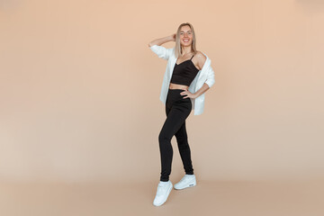 Full length of smiling young woman with freckles posing in white blazer and black clothes with carefree upbeat face expression. Indoor studio shot on beige background