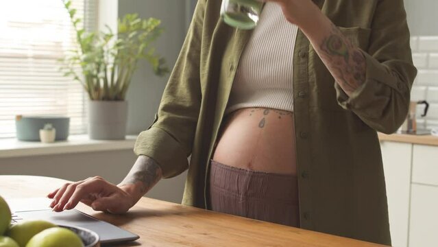 Cropped shot of tattooed woman expecting baby working on laptop from home drinking healthy green smoothie