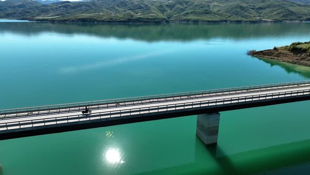 Biker riding on a long empty bridge overlooking a blue lake on a sunny day areal shot 4K 60 fps