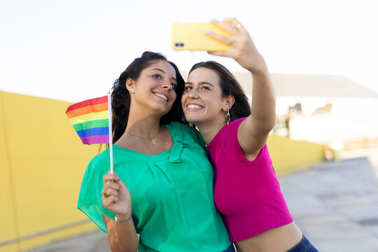 A beautiful lesbian young couple embraces and holds a rainbow flag. Girls taking selfie photo