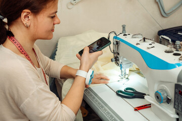 Caucasian woman tailor sews on a sewing machine in a home workshop and records the broadcast on a smartphone.Small business.Blogging concept.
