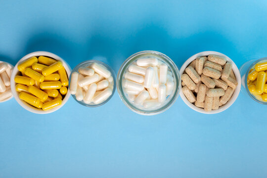 Nutritional Food Supplements, Vitamins And Minerals Such As Vitamin D, Vitamin C, Vitamin E, Vitamin B, Calcium, Zinc, Magnesium In Jars From Above On Light Blue Background