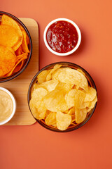 Tasty potato chips in plates and sauces on a wooden desk on orange background from above. Snack time.