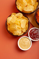 Tasty potato chips in plates and sauces on a wooden desk on orange background from above. Snack time.