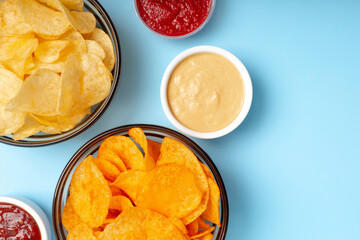 Tasty crispy potato chips in dark transparent bowls with different sauces on light blue background.