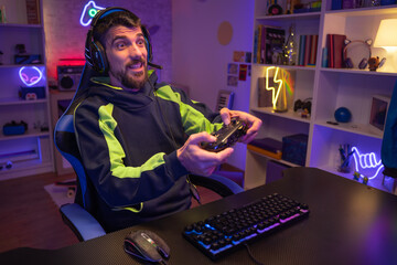 Happy guy gamer wear headphone play video game colorful neon lights computer in living room. 