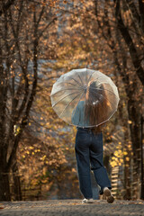 Brunette with long hair in blue sweater and pants stands with back under transparent umbrella in autumn park