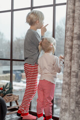 Little boy and girl in red striped pants peek out large window. Brother and sister on Christmas Eve...