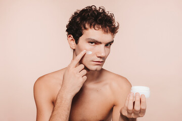 Fototapeta premium Close up portrait of attractive young man with healthy skin, applies cream moisturizer for anti wrinkle or anti aging, cares of body, poses naked or shirtless, isolated over beige background.