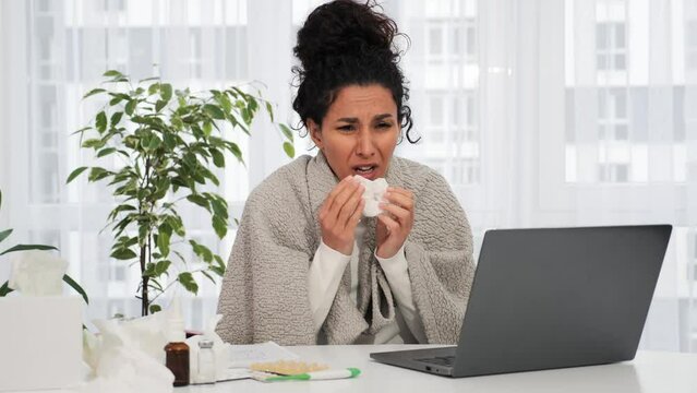 Young latin hispanic woman having allergy. Cough, sneezing, runny nose. Woman working remotely from home with laptop coughs and wipes her nose with napkin. Cold, flu, coronavirus, covid-19 concept.
