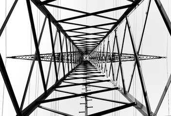 High voltage pylon for energy supply with power cables seen from underneath in frog perspective with vanishing point. Contrasting metal structure with symmetrical design, black and white greyscale.
