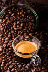 in the foreground a glass cup with espresso coffee. The cup is surrounded by roasted coffee beans.