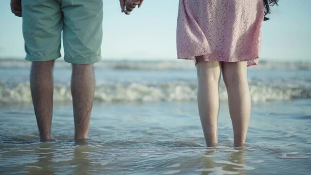 Rear shot of a couple holding hand in beach with waves hitting their legs