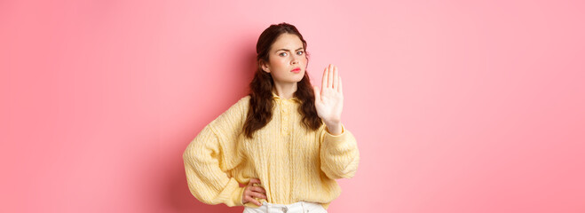 Angry and bossy young woman frowning, looking serious, showing block stop gesture, stretch out hand to say no, refuse something bad, forbid action, standing against pink background