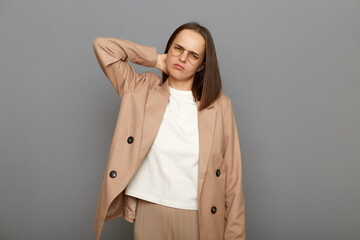 Indoor shot of unhealthy sick upset woman with dark hair wearing jacket posing against gray wall, having neck pain or ache, standing and holding her painful neck.