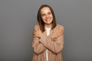 I love myself. Pleased satisfied female wearing beige jacket standing isolated over gray background, hugging herself and smiling, feeling comfortable and fulfilled, narcissistic egoistic person.