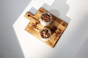 Yoghurts in glass jars with granola, chocolate and nuts on a wooden plank on a white background. The concept of healthy eating. Flat lay