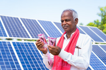 Happy farmer counting money or currency notes in front of solar panel at farmland - concept of loan...
