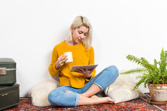 Woman having a tea, sitting on a floor in an apartment, writing down notes, opened laptop in front of her. Home office concept.