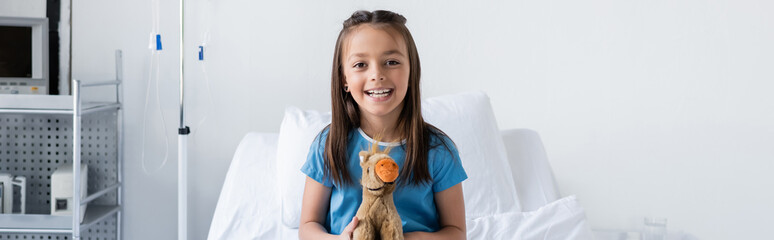 Cheerful kid holding soft toy and looking at camera in clinic, banner.