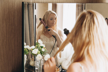Woman curls her hair on curling iron in front of mirror at dressing table. Preparing for party