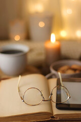 Bowl of cookies, cup of tea or coffee, chocolate, spices, knitted blanket, books, glasses and candle on the table. Cozy hygge atmosphere at home. Selective focus.