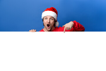 Photo of surprised man wear red clothes and x-mas hat hold empty space poster ad isolated on blue color background, Christmas, new year concept