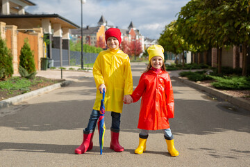 Two charming children, a boy and a girl, in waterproof raincoats and rubber boots with an umbrella.