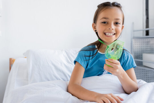 Cheerful kid holding oxygen mask and looking at camera on bed in clinic.