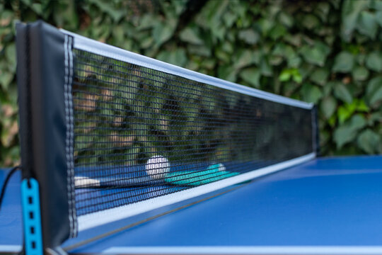 
Ping pong rackets and balls on a blue table with a net, outdoors, close-up view, diffuse photography with selective focus.