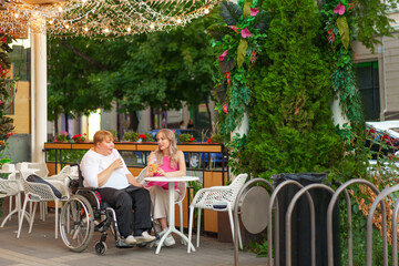 Young daughter and her mother in wheelchair sitting at the table in cafe with drinks and having fun