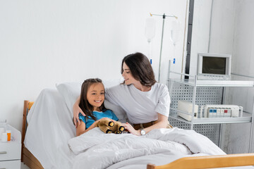 Obraz premium Positive woman hugging daughter with toy on clinic bed.