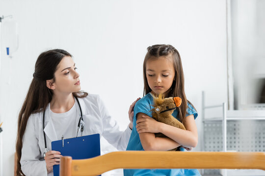 Pediatrician holding clipboard and calming upset kid with soft toy in hospital ward.