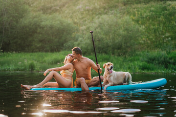 Young couple in love sitting on pad board with dog on river at summertime during vacations kissing. Weekend and outdoor leisure activities. People and domestic animals. Husband kissing wife.