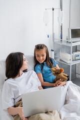 Smiling child in patient gown looking at laptop near mom on hospital bed.
