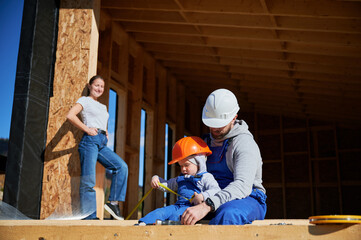 Father, mother and son building wooden frame house in the Scandinavian style barnhouse. Toddler boy helping his daddy, while woman looking for them on construction site. Carpentry, family concept.