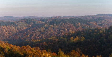 Hills overgrown with lush colorful forest in autumn, sunlit by evening sun in haze, autumn landscape