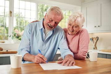 Retired Senior Couple Sitting In Kitchen At Home Signing Financial Document - obrazy, fototapety, plakaty
