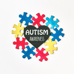 Autism campaign symbols. Colorful puzzle pieces around blackboard stone heart with words Autism Awareness written in chalk. Flat lay, top view, square composition on off white background.