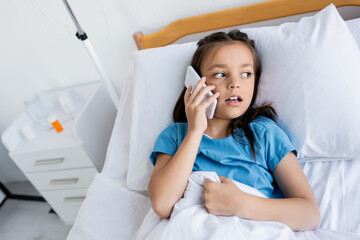 Ill child talking on smartphone while lying on bed in clinic.