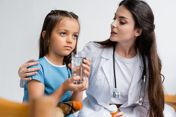Doctor holding pills and talking to girl with glass of water in hospital ward.