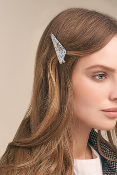 Cropped shot a girl with blonde hair. The lady's hair is fixed with a glittering hair clip in the shape of a triangle. The woman in a white blouse and a plaid jacket is posing on the grey backdrop.