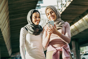 Two young Muslim females covered with Islamic hijab relaxing after jogging at the city street. They're using smartphone and talking.	
