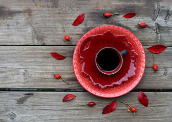 En kopp svart kaffe, koppen står på två röda tallrikar. Runt om finns röda löv och nypon. Bilden är tagen på trä.  - obrazy, fototapety, plakaty