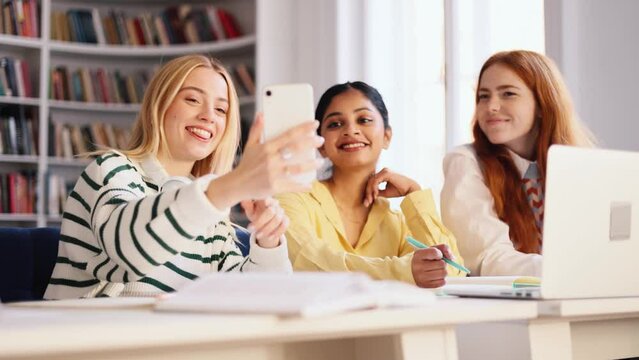 Charming blond girl holding smartphone and show funny pictures to her interracial classmates at university library after lessons. Friends at university.