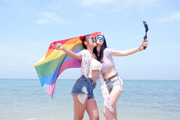 A lesbian couple holding a rainbow flag of the LGBT symbol and holding a phone. Take a selfie at the beach LGBT Concepts.