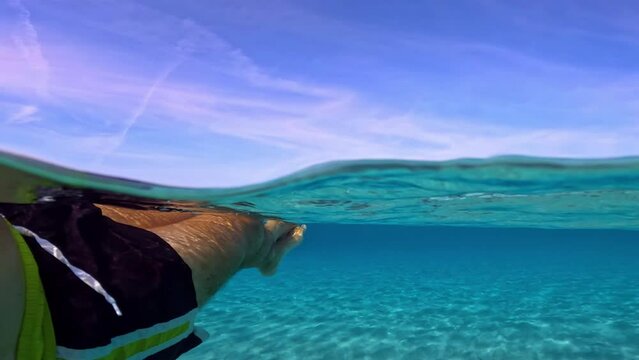 Personal perspective of male crossed legs and feet relaxing while floating on turquoise sea water of Saleccia tropical beach in Corsica island, France. Half underwater slow motion scene