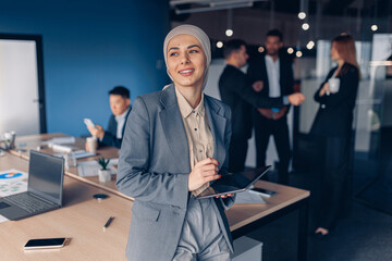 Smiling muslim businesswoman in hijab working on digital tablet while standing in modern office