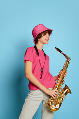 Fototapeta na wymiar Portrait of cheerful teen girl with curly brown short hair posing in pink panama and playing saxophone isolated over blue background